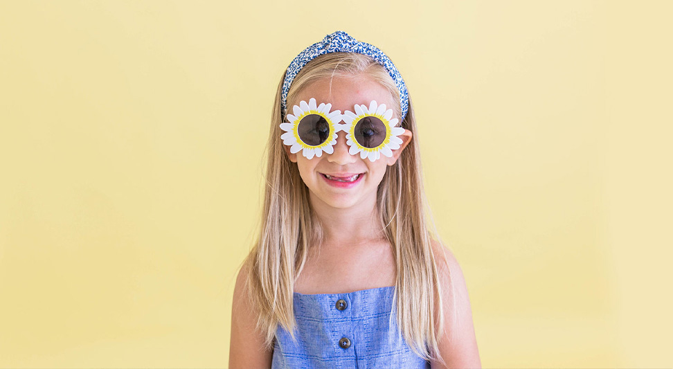 nJoy Vision OKC LASIK Blog UV Safety Awareness Month image of cut young girl with daisy sunglasses against a yellow background